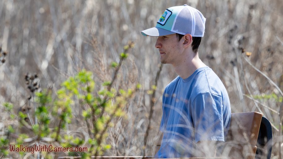 A person wearing a cap is shown in profile against a woodland background. Thin branches and fresh leaves cross the soft foreground.