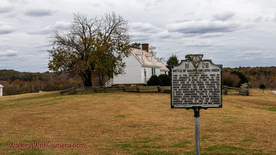 A historic marker stands in front of a small white building and mature trees. Open grass and a cloudy sky frame the scene.