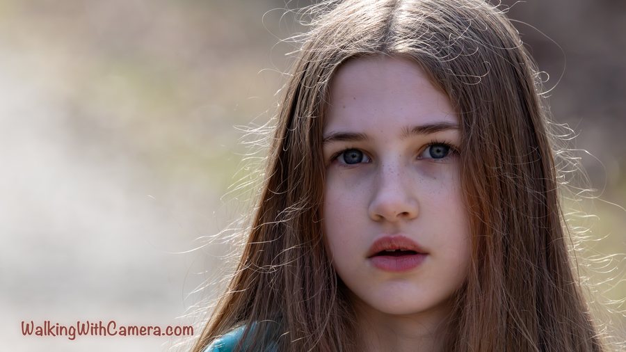 A close portrait shows long brown hair and a direct gaze. The face is sharp against a softly blurred outdoor background.