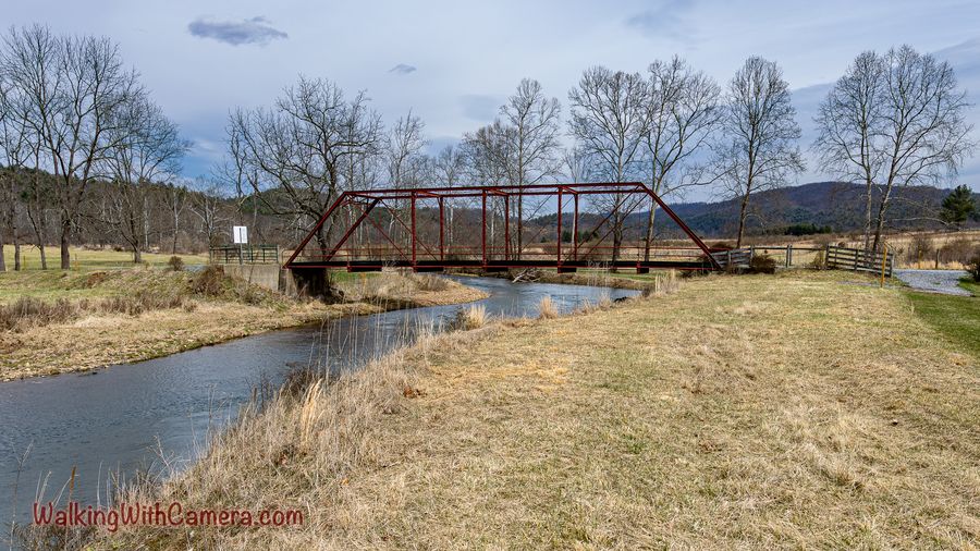 A red-brown metal footbridge crosses a narrow stream amid open grass and bare trees.