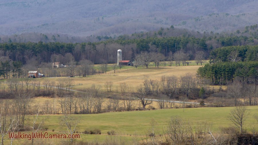 Fields, bare trees, farm buildings, and a tall silo spread below distant wooded hills.