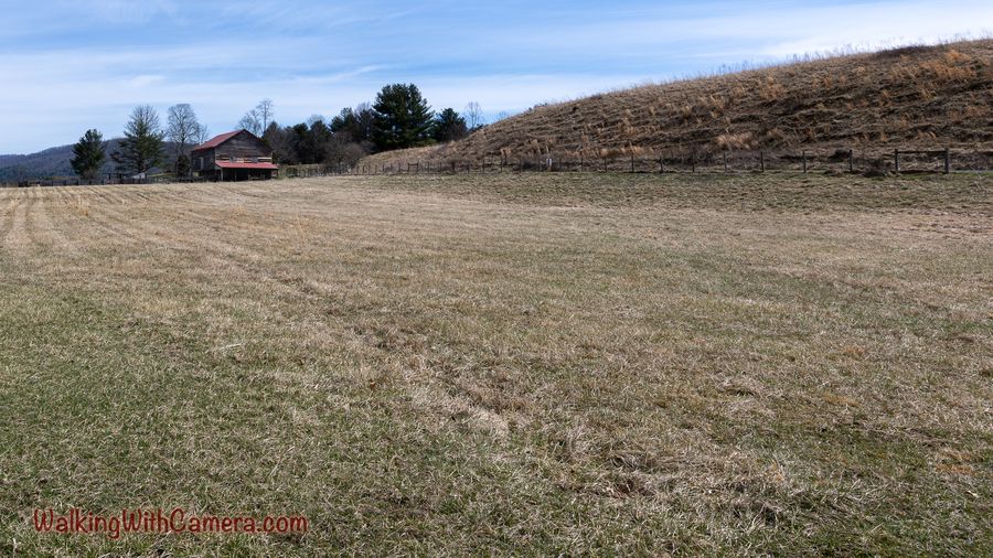 A broad grassy field slopes toward a barn, fence, and distant hills.