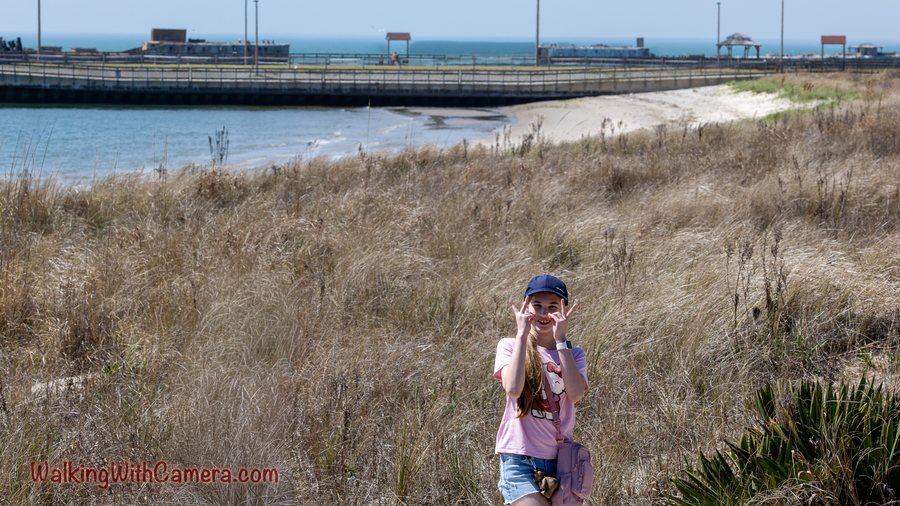 A person poses among shoreline grass with water and a long structure behind them.