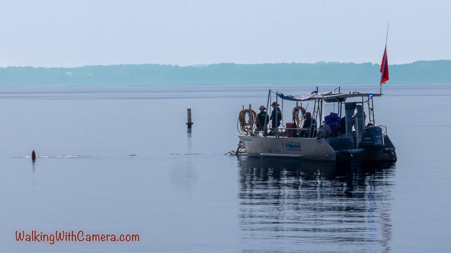A small boat sits on calm water with a red flag raised. Its structure and passengers are visible above a clear reflection, with a low shoreline in the distance.