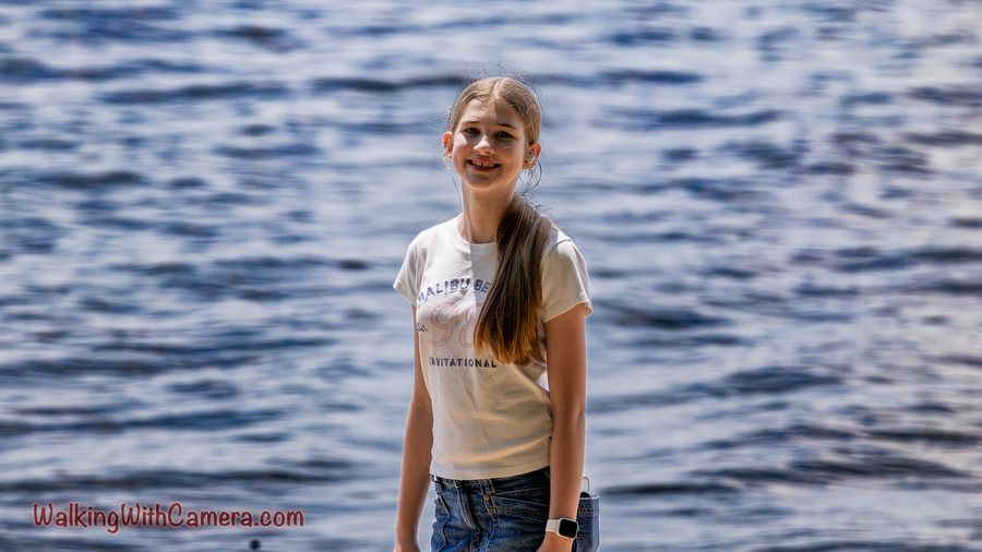 A person stands beside water with a relaxed smile. Ripples fill the blue background, and a light-colored shirt contrasts with the darker water.