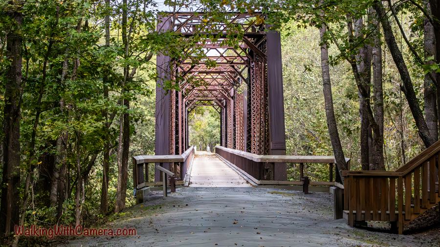 A bridge framed by rust-colored metal trusses runs through green woodland. Repeating beams draw the eye toward the bright opening at the far end.