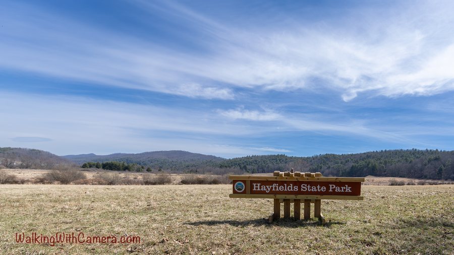 A wooden state-park sign stands in an open field below distant hills and a blue sky.