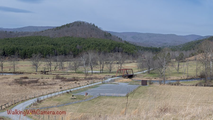 A stream and footbridge sit below open fields, bare trees, and forested hills.