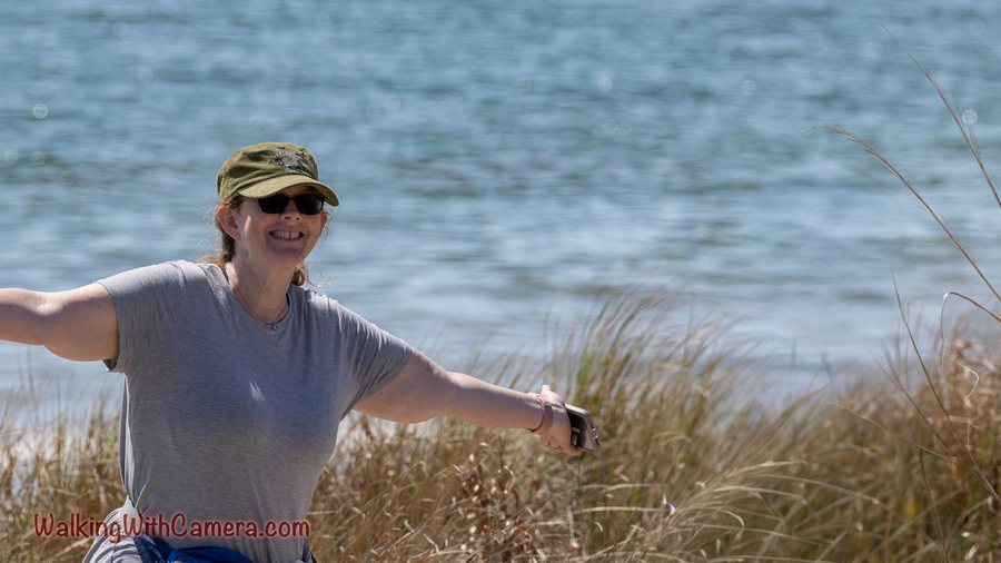A smiling person wearing a cap and sunglasses stretches both arms beside the water.