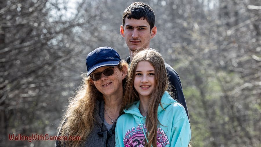 Three people stand together for an outdoor portrait. Their faces fill the center of the frame against a softly blurred woodland background.