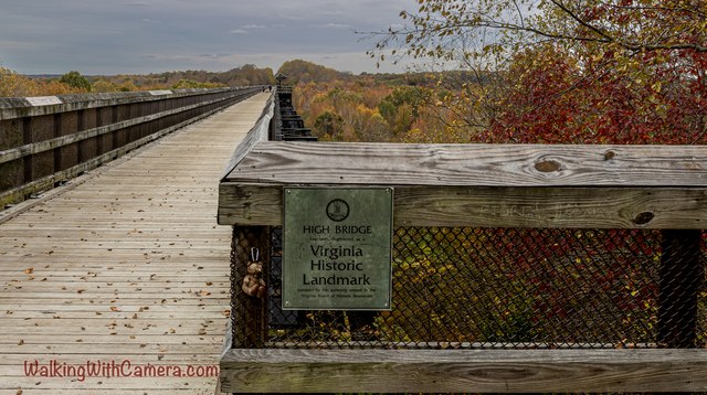 A plaque reading High Bridge Virginia Historic Landmark is fixed to a wooden railing. The long bridge deck stretches behind it above autumn-colored trees.