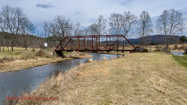 A red-brown metal footbridge crosses a narrow stream amid open grass and bare trees.