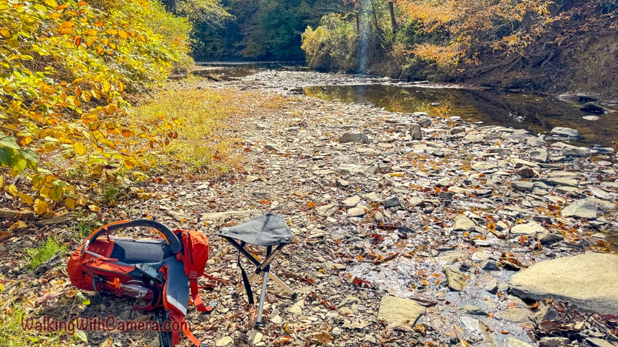 A shallow creek runs over a broad bed of stones and fallen leaves; a backpack and folding stool sit on the sunny bank.