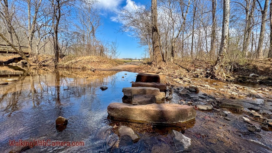 A shallow creek winds through leafless woods. Large flat rocks form a stepping-stone crossing, with a wooden footbridge visible at the left and trees reflected in the water.