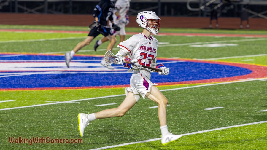 A lacrosse player wearing a white Red Wolves uniform and helmet runs across a green field with a stick in hand; other players and a colorful midfield logo are visible behind.