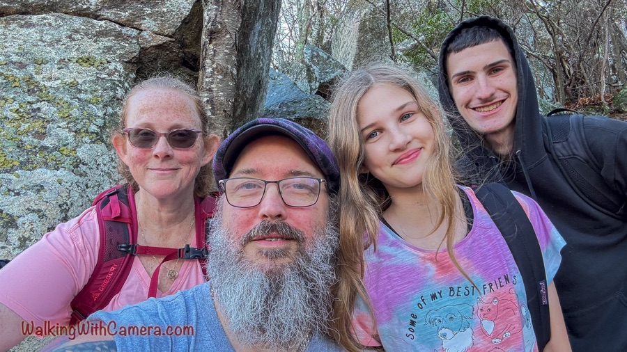 A close group selfie shows four people smiling in hiking clothes in front of a large lichen-covered rock and trees.