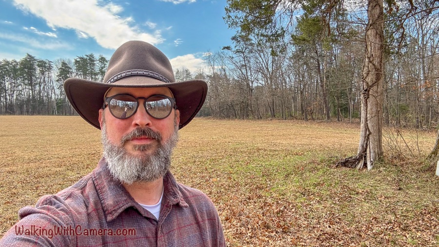 A bearded man wearing a brown cowboy hat and sunglasses takes a selfie in front of a grassy field and leafless woods.