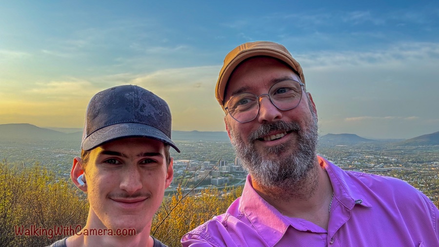 Two people smile for a summit selfie with a city and distant ridges behind them in warm evening light.