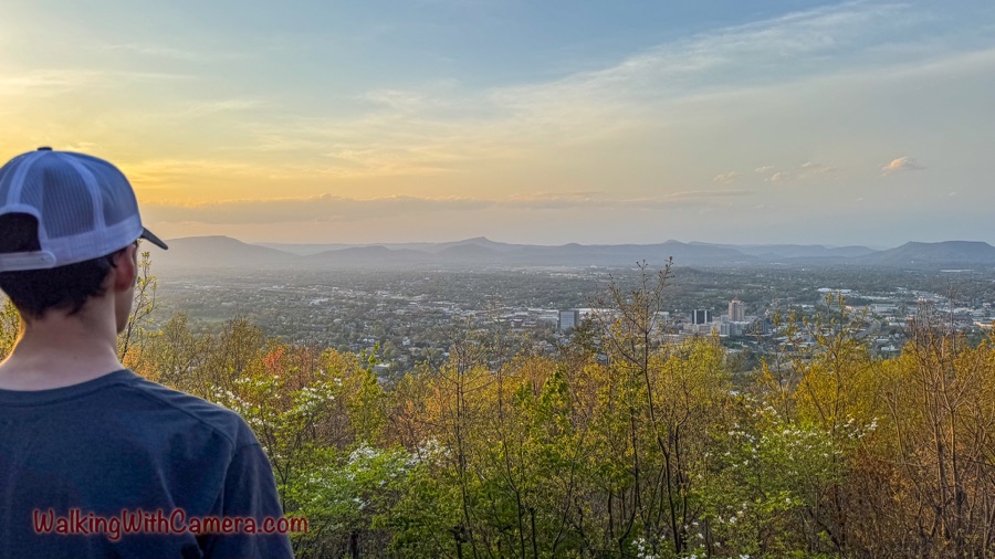 A hiker seen from behind looks out over a city and layered mountain ridges beneath a softly lit evening sky.