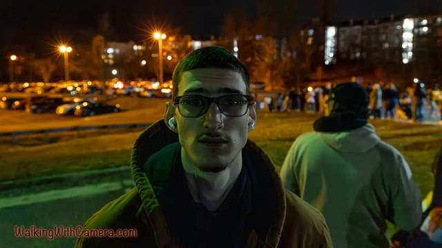 A person wearing glasses faces the camera beneath outdoor evening lights.