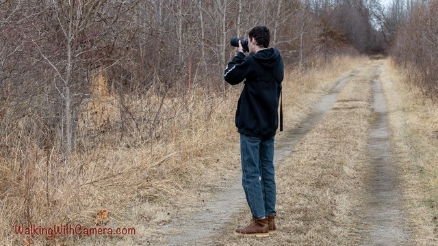 A person holds a camera toward the trees while standing on a quiet gravel track.