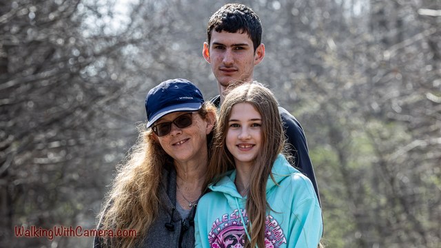 Three people stand together for an outdoor portrait. Their faces fill the center of the frame against a softly blurred woodland background.