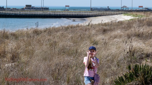 A person poses among shoreline grass with water and a long structure behind them.