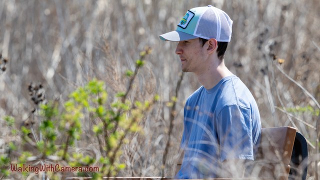 A person wearing a cap is shown in profile against a woodland background. Thin branches and fresh leaves cross the soft foreground.