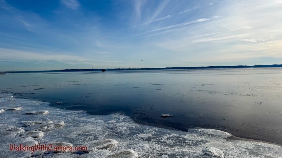 Ice along the shore meets dark, calm water beneath a wide blue sky and distant horizon.