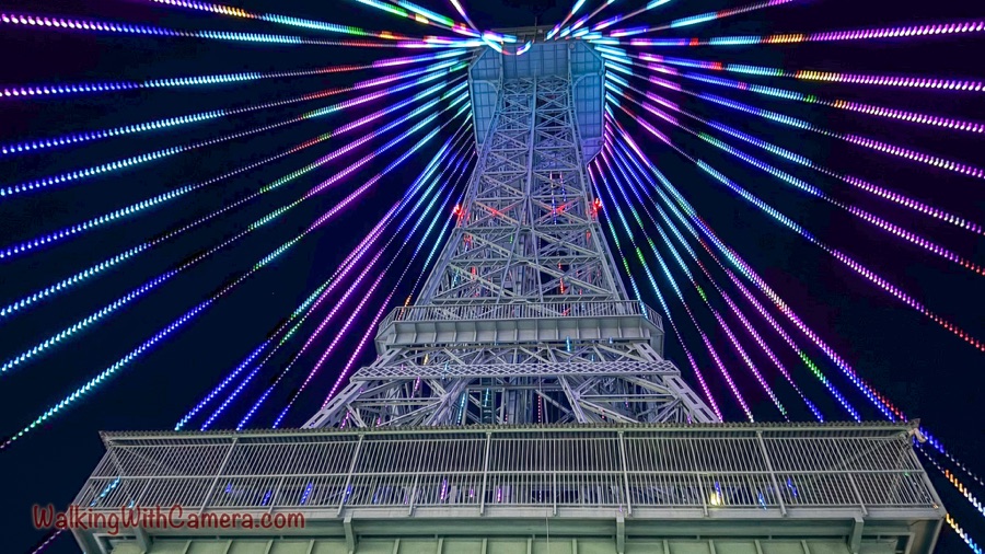 A tall metal tower viewed from below is surrounded by colorful lines of lights against a dark night sky.